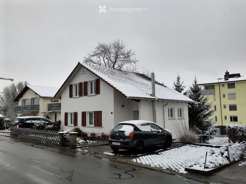 Two-story house with white exterior, red shutters, snow-covered roof, and a driveway with parked cars.
