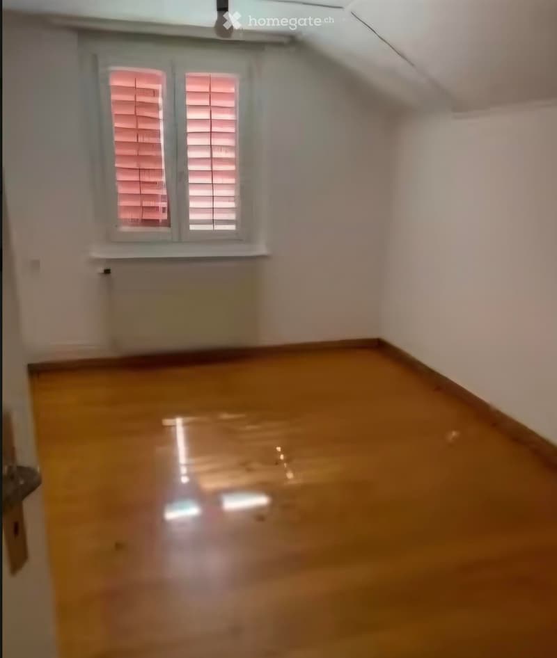 Empty room with wooden flooring and white walls, featuring a window with blinds and a door on the left side.