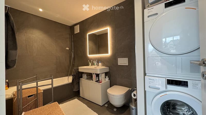 Modern bathroom with dark grey tiles, a white sink with a built-in mirror, a toilet, a bathtub, a small cabinet, and a washing machine/dryer unit.
