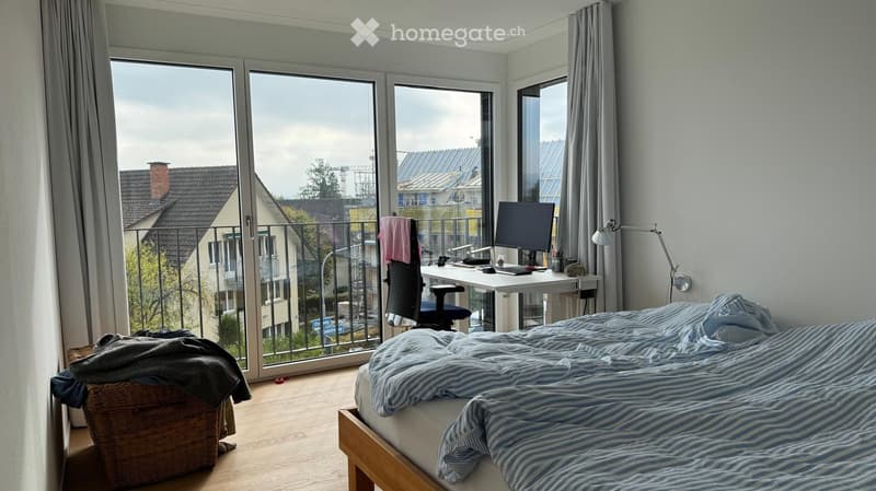 Modern bedroom with a large window and balcony access, featuring a bed with striped bedding, a desk with a computer, and a wicker basket on the floor.