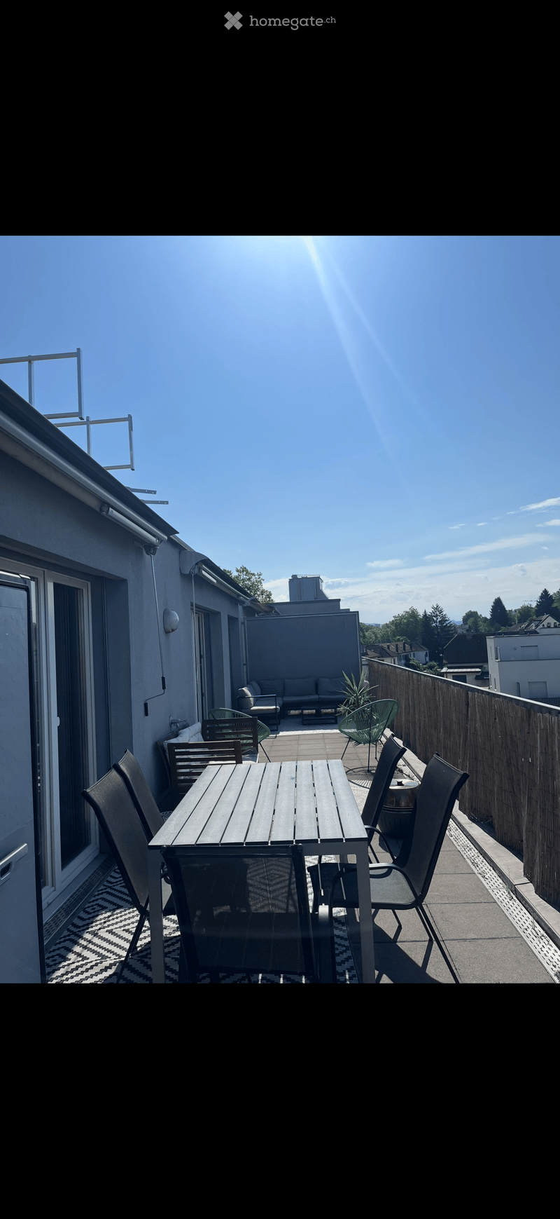 Balcony with wooden table, chairs, sofa, and a small plant, surrounded by a wooden fence and clear blue sky.