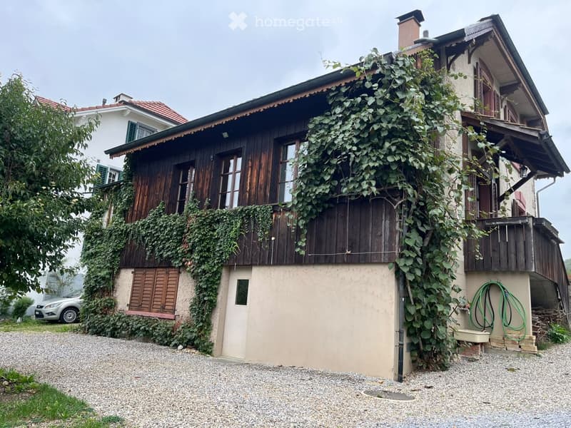 Two-story house with wooden facade, ivy-covered walls, and a gravel driveway. Features include a chimney, multiple windows, and a small garden area with a hose reel.