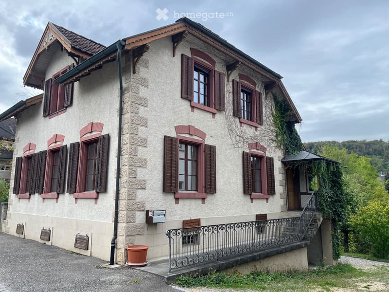 Two-story house with beige stucco walls, red shutters, and a tiled roof. Features a small balcony with ornate metal railing and a potted plant on the ground.