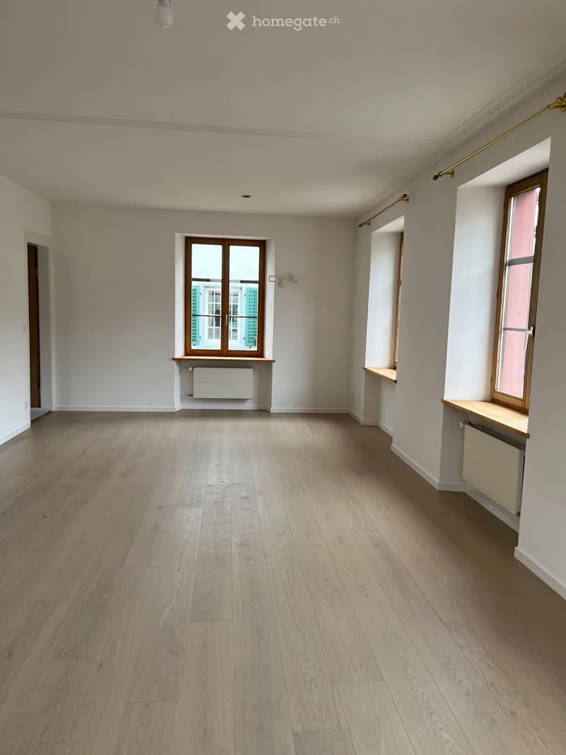 Empty room with light wooden flooring, white walls, and large windows with wooden frames. Radiators are visible beneath the windows.