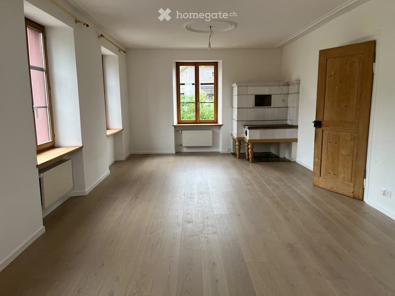 Empty room with wooden flooring, white walls, and large windows. Features include a built-in bench, radiator, and a small cabinet with a sink.