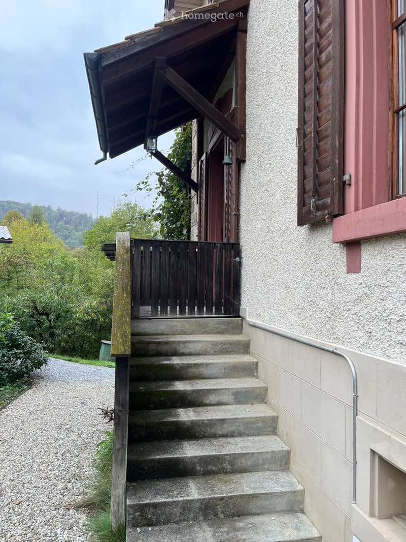 A two-story house with a wooden porch, concrete steps leading up to the entrance, and a gravel driveway.