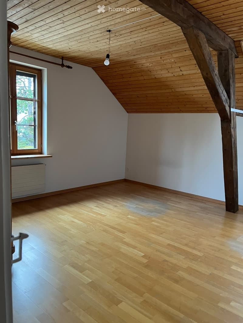 Empty room with wooden flooring, slanted wooden ceiling, exposed wooden beam, single window with white frame, radiator, and pendant light fixture.