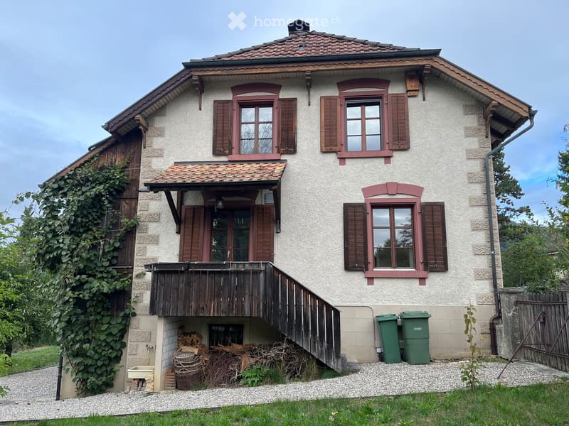 Two-story house with a tiled roof, wooden shutters, and a covered entrance. The house has a stone facade and a wooden balcony. There is a small garden area with a stack of firewood and two green trash bins.
