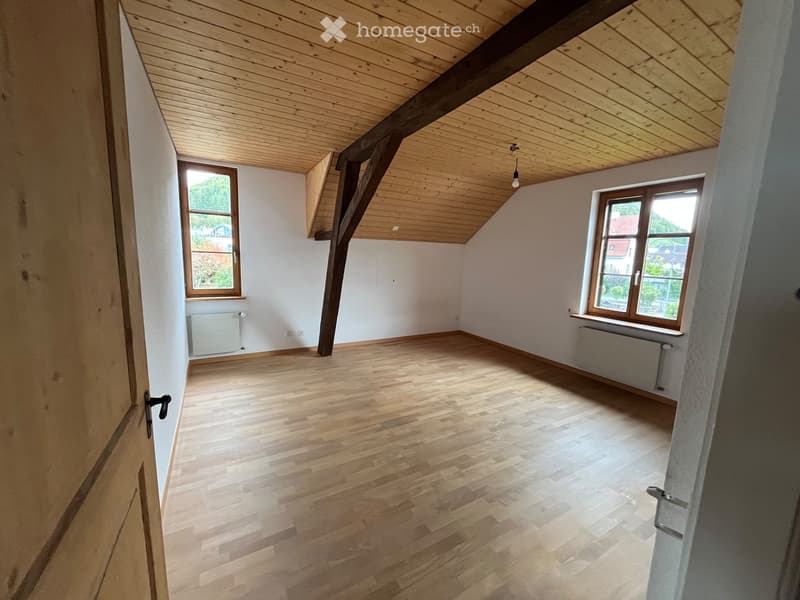Empty attic room with wooden floors, white walls, exposed wooden beams, and two windows offering a view of the outside.