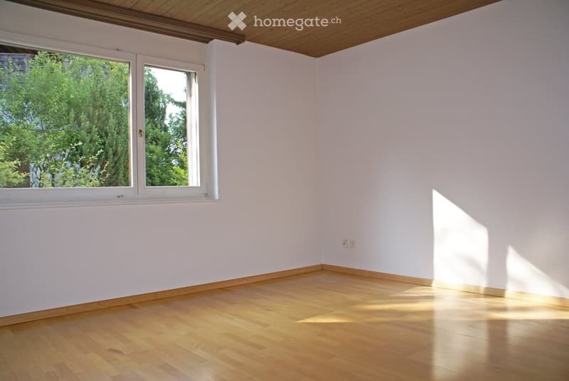 Empty room with light wooden flooring, white walls, and large windows allowing natural light to enter. The ceiling is wooden, and there are visible electrical outlets on the wall.