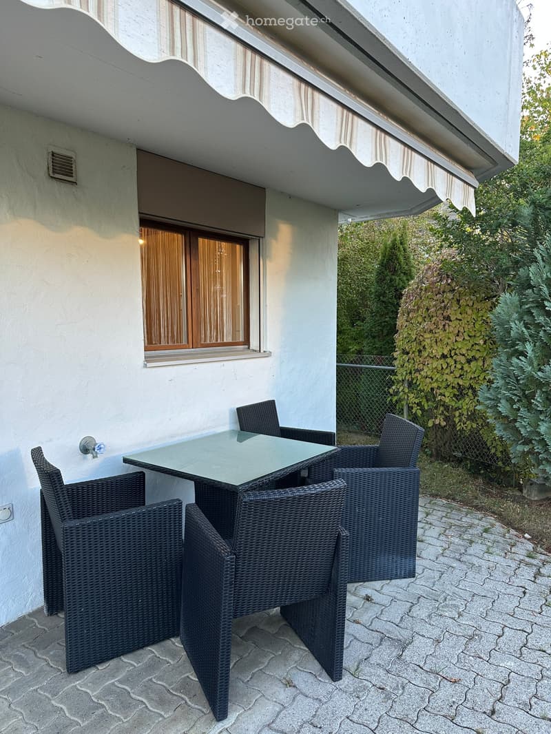 Balcony with a glass table and four wicker chairs, situated next to a building with a covered awning and a window with curtains.