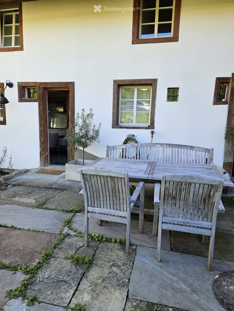 Outdoor patio with stone flooring, wooden table, chairs, and bench, surrounded by small plants and greenery.