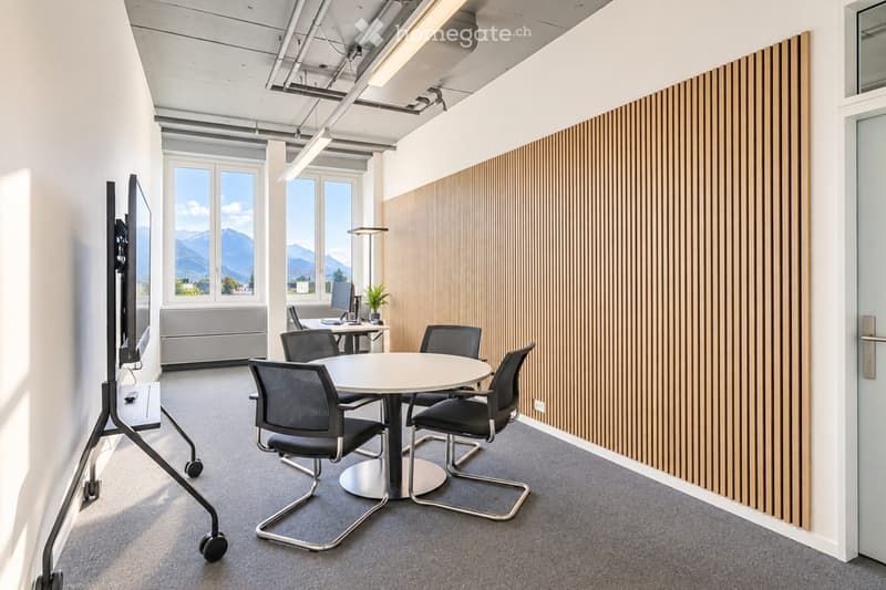 Modern office meeting room with a round table, chairs, a large screen, and a computer setup, overlooking a scenic mountain view.
