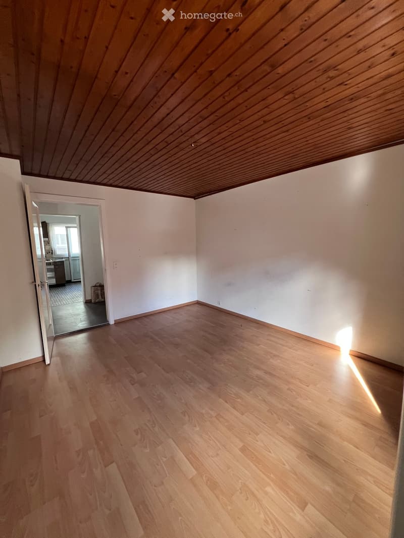 Empty room with light wooden flooring and white walls, featuring a slanted wooden ceiling and an open doorway leading to another room.