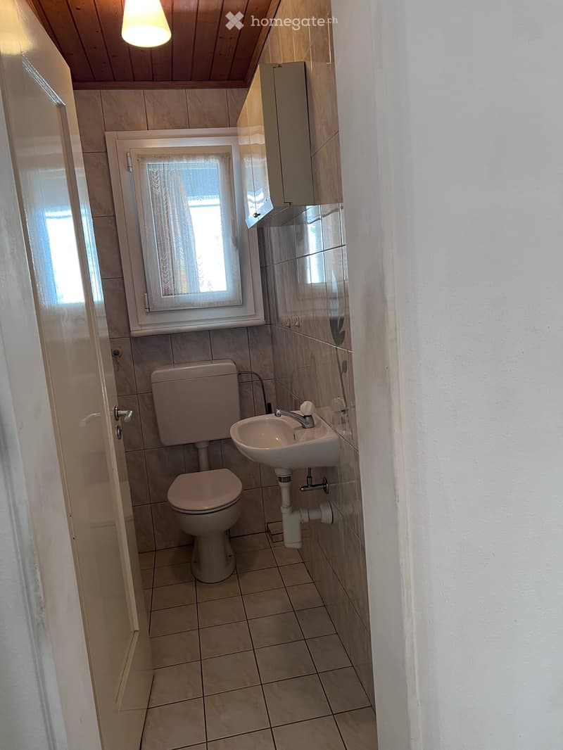 Small bathroom with beige tiles, a toilet, a sink with a faucet, a mirror, and a wooden cabinet above the sink. The window has a white frame and a curtain.