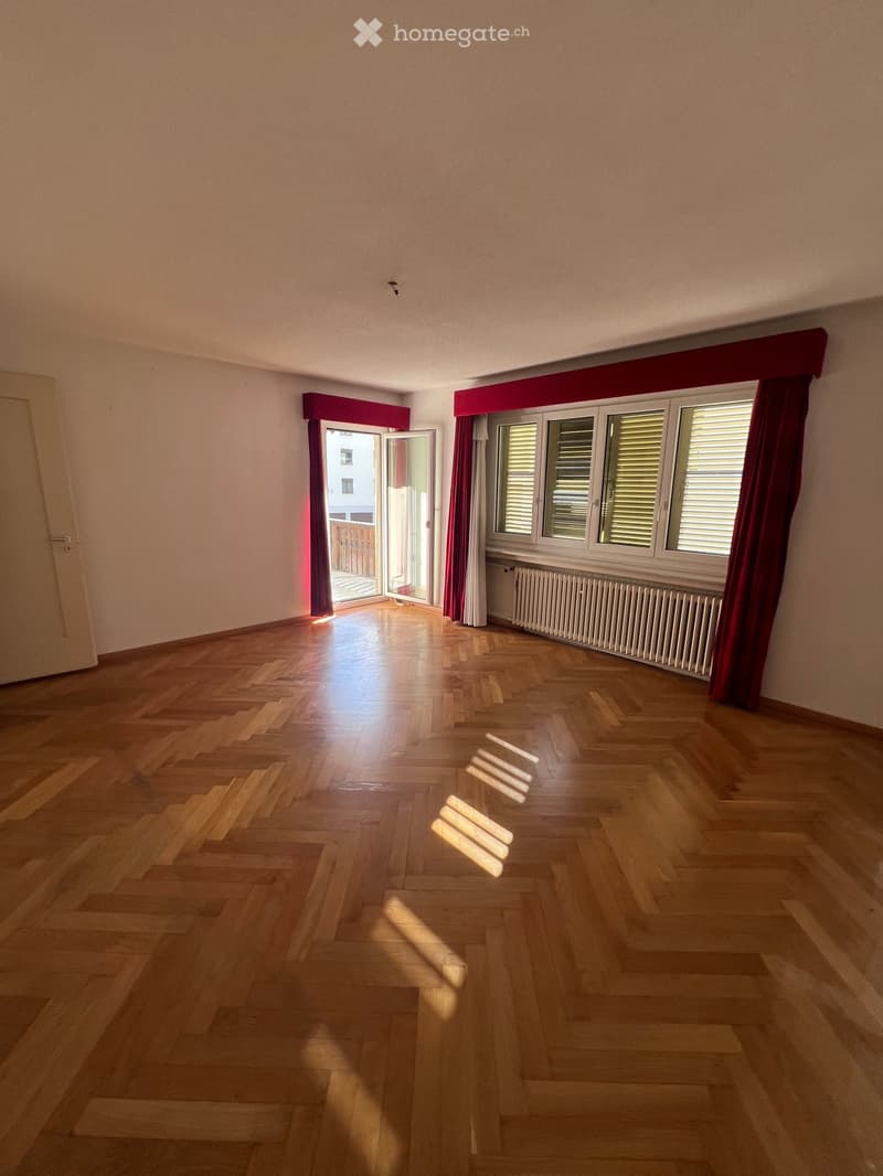 Empty room with wooden parquet flooring, large windows with red curtains, and an open door leading to a balcony.