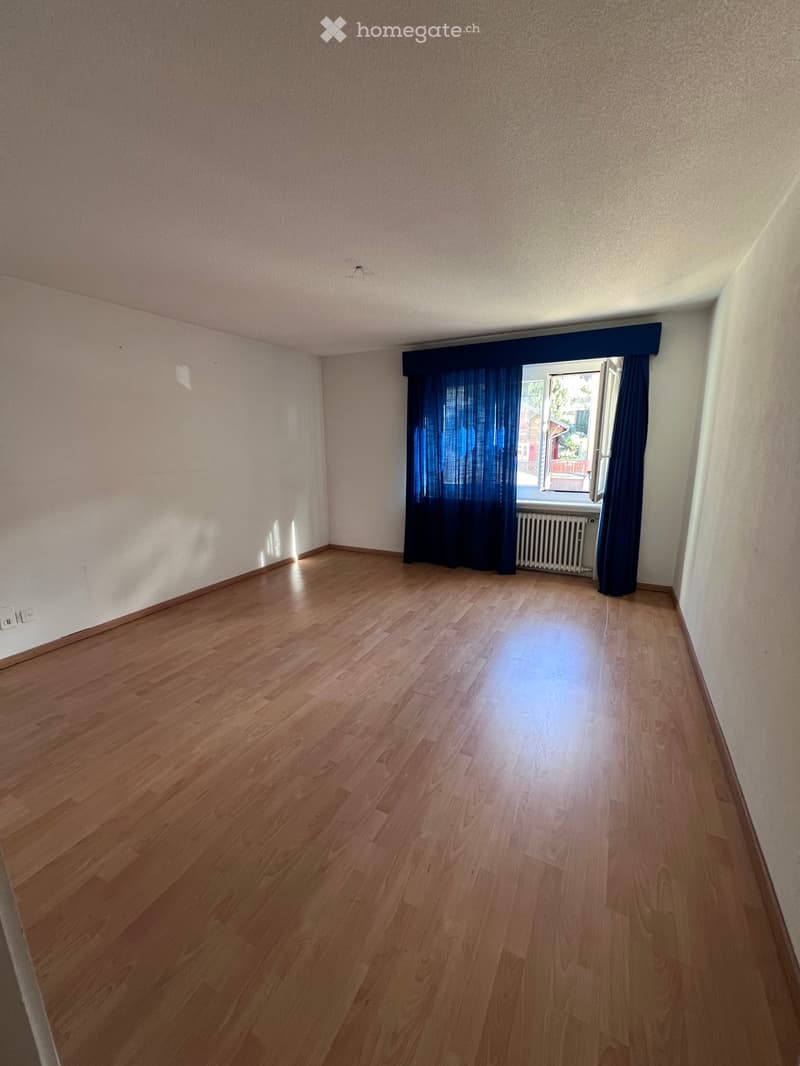 Empty room with wooden flooring, white walls, and a window with blue curtains. No furniture or equipment is present.