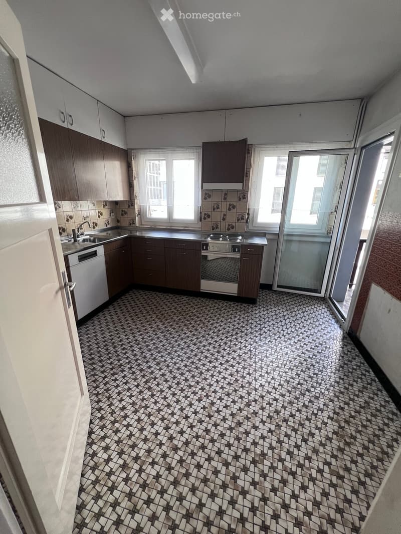 Modern kitchen with dark wooden cabinets, white appliances, and a patterned tile floor. Features a sink, dishwasher, stove, and oven, with large windows and a sliding glass door leading outside.