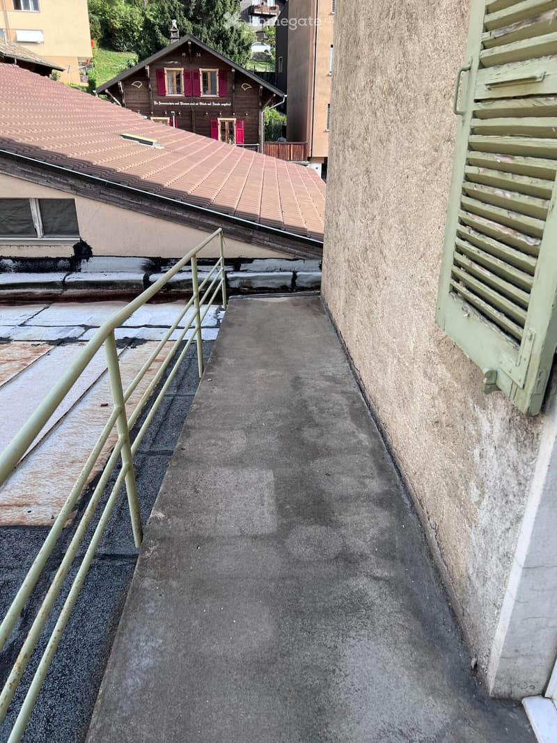 A narrow concrete pathway with a metal railing on one side, leading between buildings with visible steps and a wooden shutter on the right.