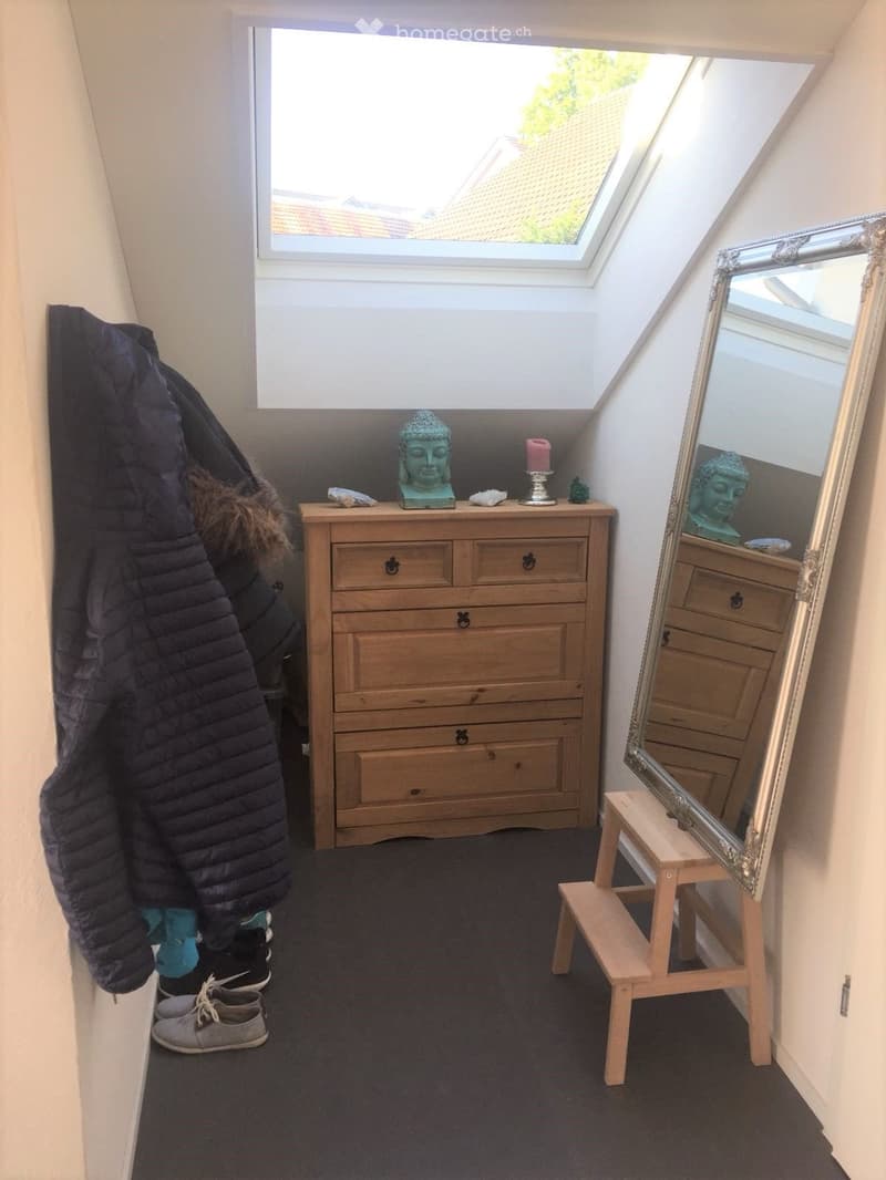 A small, furnished room with a wooden dresser, a large mirror on a stool, a statue, a candle, and a coat hanging on the wall. The room has skylight windows and a dark carpeted floor.