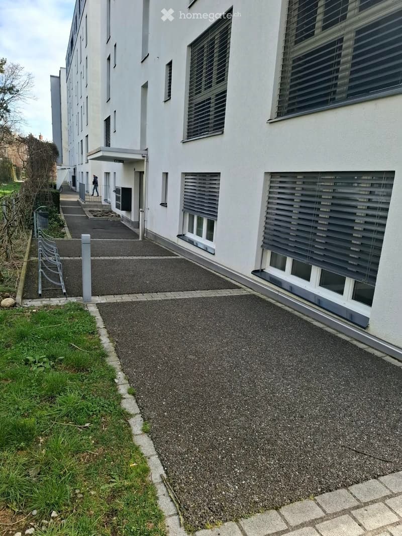 Modern multi-story building with white facade, large windows, and a covered entrance area. The ground level features a paved walkway and a small grassy area.