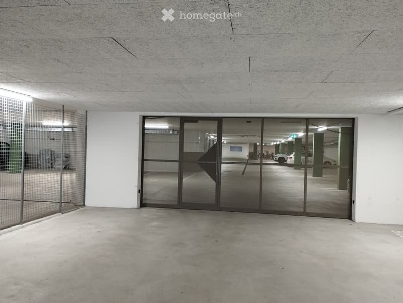 Empty underground parking space with concrete floors and walls, featuring a large glass door leading to another parking area.