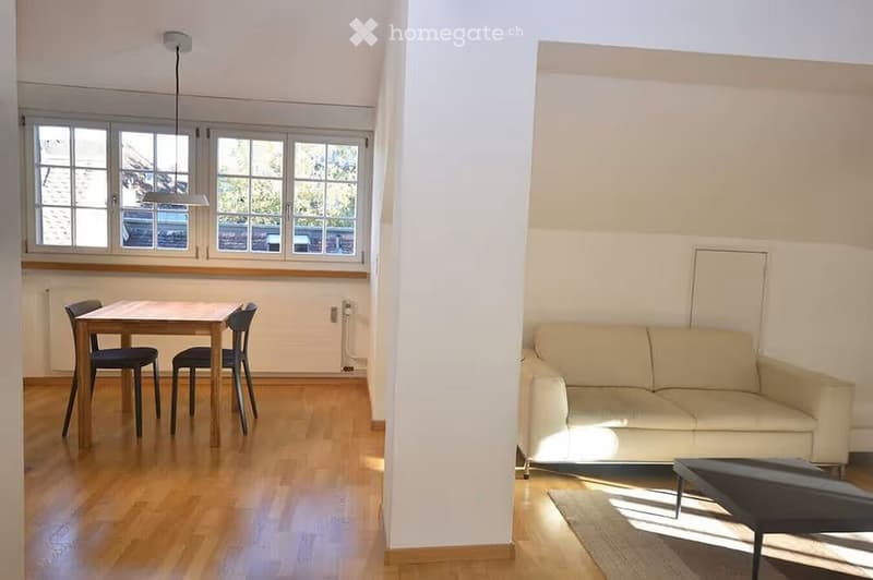 Bright, open-plan living and dining area with wooden flooring, large windows, a cream sofa, coffee table, and a dining table with chairs.