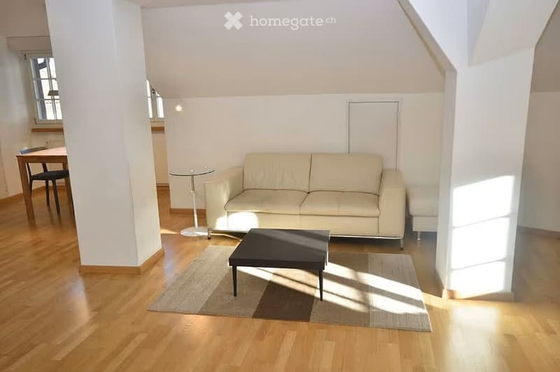 Bright living room with wooden flooring, white walls, and large windows allowing natural light. Features a beige sofa, black coffee table, small round glass table, and a dining area with a table and chairs.