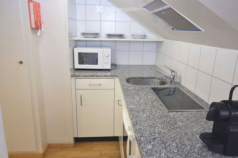 Compact kitchen with granite countertops, white cabinets, microwave, sink, and stove. Plates are stacked on a shelf above the microwave.