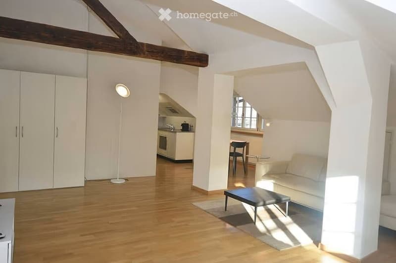 Bright, open-plan living and dining area with wooden flooring, white walls, and exposed wooden beams. Features a white sofa, a small black stool, a round glass table, a chair, a floor lamp, and a built-in kitchen with an oven and sink.