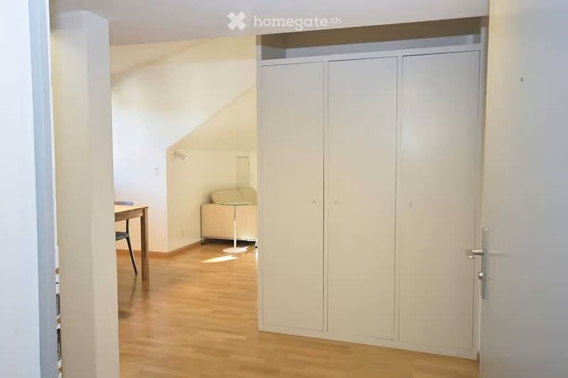 A small, well-lit room with wooden flooring, a built-in closet, a small table with a chair, and a lamp near the corner.