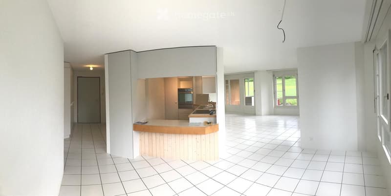 Empty room with white tiled flooring, white walls, and a small kitchen area with a counter and built-in appliances visible in the background.