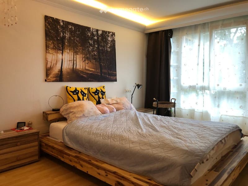 Cozy bedroom with a wooden bed frame, white bedding, nightstand, and a large forest-themed picture on the wall.
