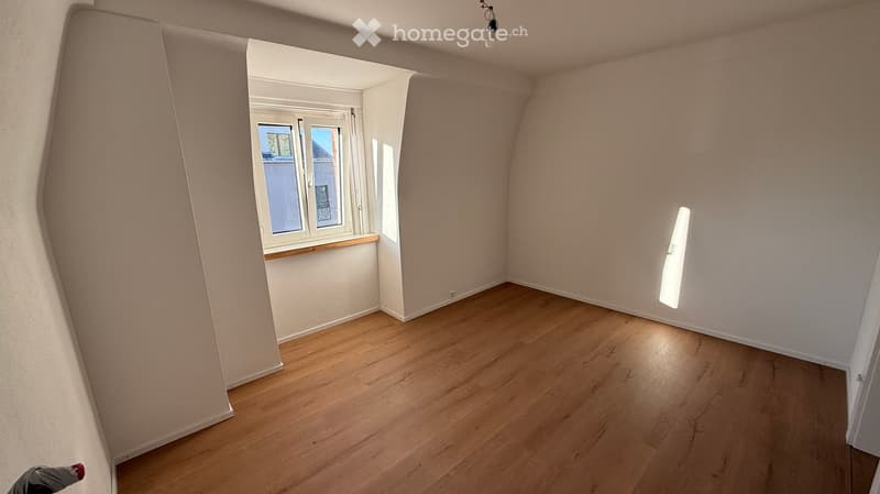 Empty room with wooden flooring, white walls, and a window with a view of the outside. The room has a slanted ceiling typical of an attic space.