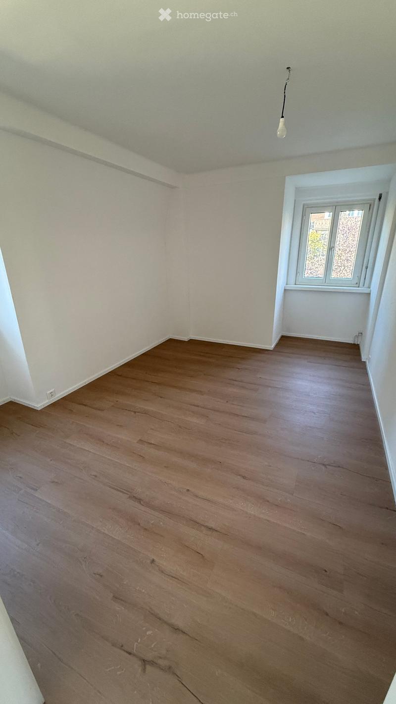 Empty room with wooden flooring and white walls, featuring a single window with a view and a hanging light fixture.