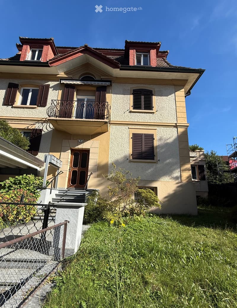 Two-story house with beige and cream exterior, multiple windows with shutters, a balcony on the second floor, and a staircase leading to the entrance.
