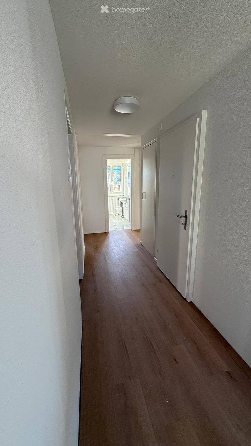 A clean, well-lit hallway with wooden flooring, white walls, and a single ceiling light fixture. Doors lead to adjacent rooms, including a visible bathroom with a toilet and sink.