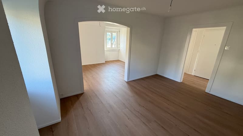 Empty room with wooden flooring and white walls, featuring an open doorway leading to another room with a window.