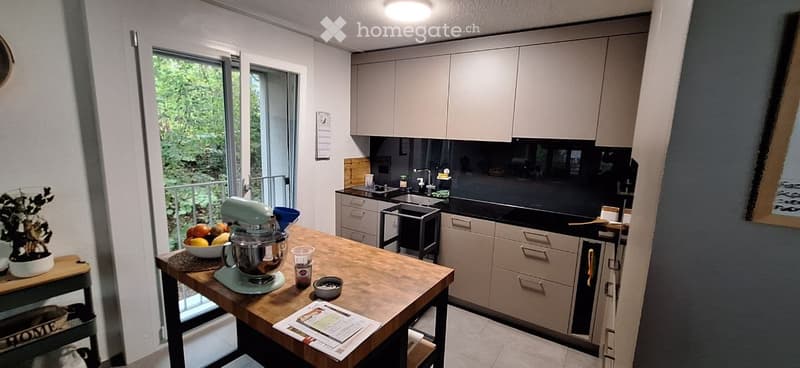 Modern kitchen with light grey cabinets, black countertops, and a wooden island. The island features a mixer, bowl, and other small items. A sliding glass door offers a view of greenery outside.