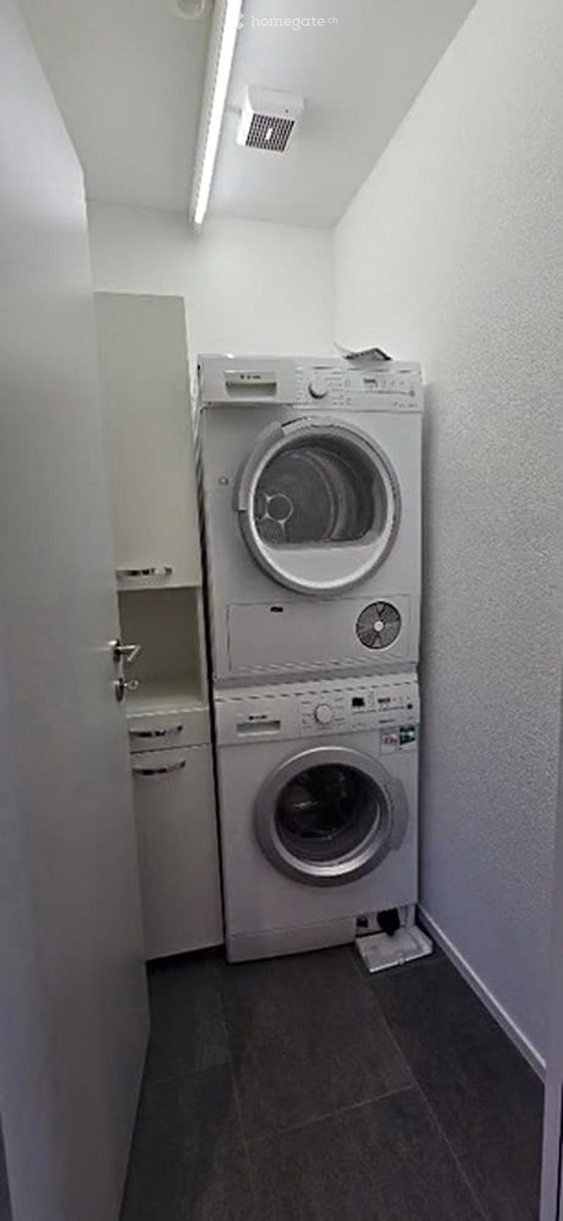 Compact bathroom with a stacked washing machine and dryer unit, white walls, and dark tiled floor.