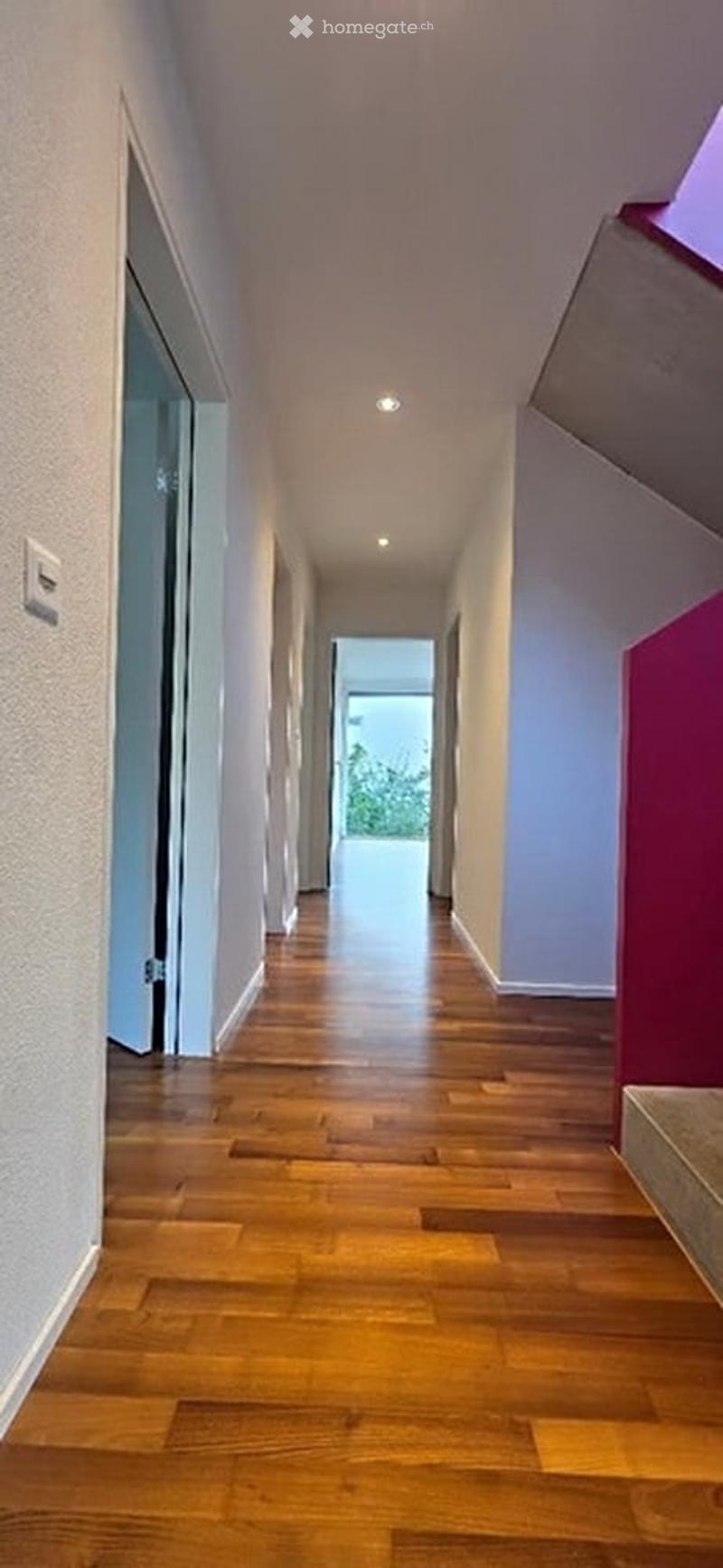 A long, narrow hallway with wooden flooring, white walls, and recessed lighting. The hallway leads to an open doorway at the end, with a glass door visible on the left side.