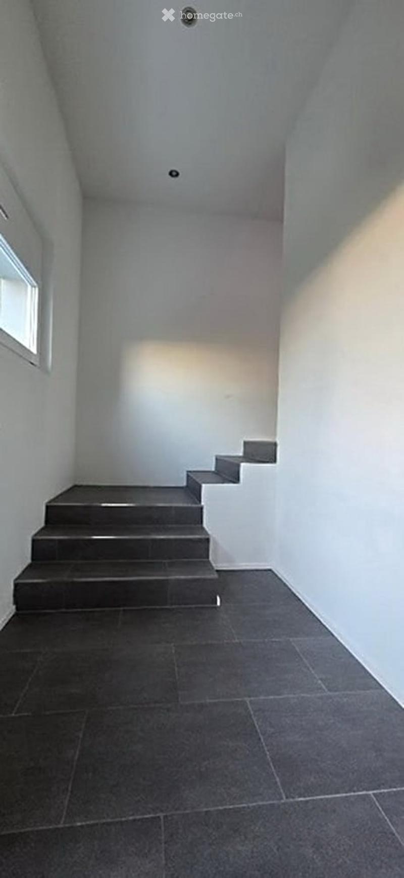 Minimalist hallway with grey tiled floor, white walls, and a small window on the left side. The ceiling has recessed lighting.