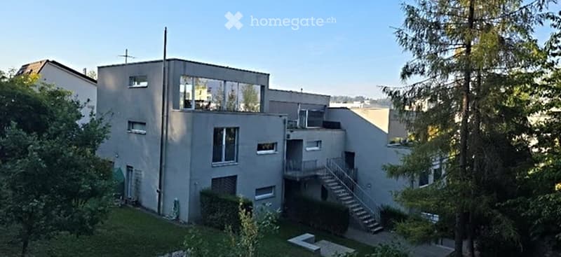 Modern two-story house with grey facade, large windows, and a staircase leading to a balcony on the upper level.