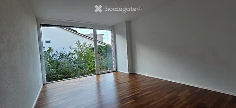 Empty room with wooden flooring and large window offering a view of greenery outside.