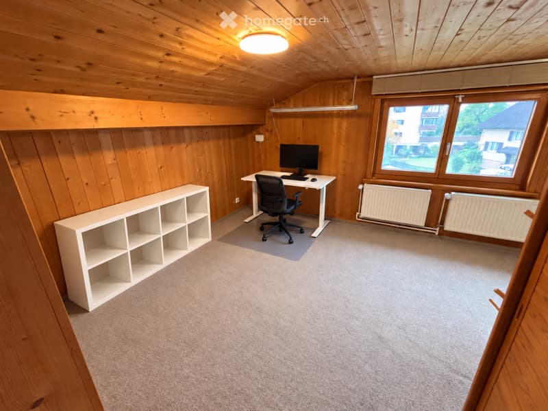 A small, well-lit room with wooden walls and ceiling, featuring a desk with a computer and chair, a white shelving unit, and large windows offering a view of the outside.