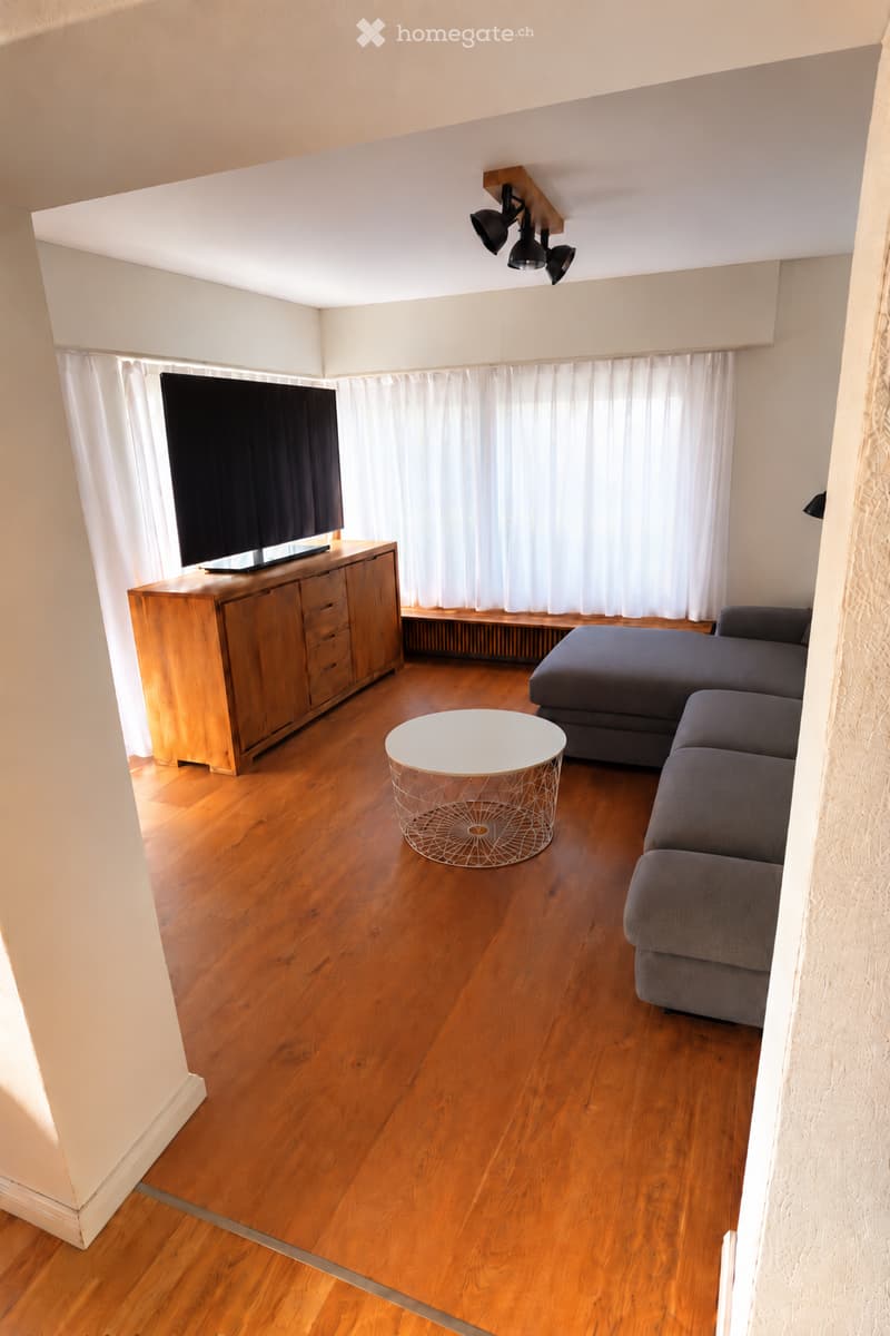 Modern living room with wooden flooring, grey sectional sofa, white coffee table, TV on wooden stand, and large windows with white curtains.