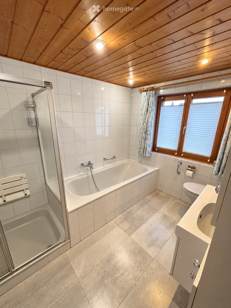 Modern bathroom with a bathtub, toilet, sink, and a glass-enclosed shower. The room features white tiled walls and a wooden ceiling with recessed lighting. There are two windows with wooden frames and a curtain on one side.
