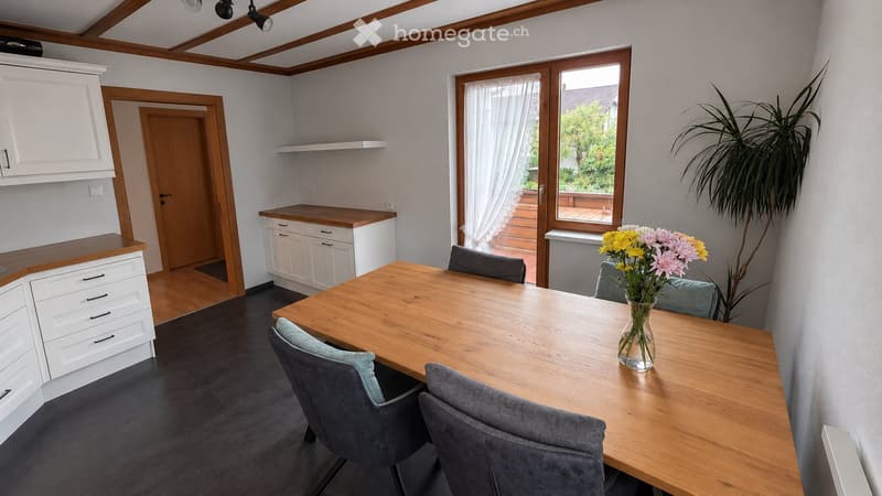 Bright kitchen with white cabinets, wooden countertops, a dining table with four chairs, and a vase of flowers on the table. Large windows with white curtains and a view of a wooden deck outside.