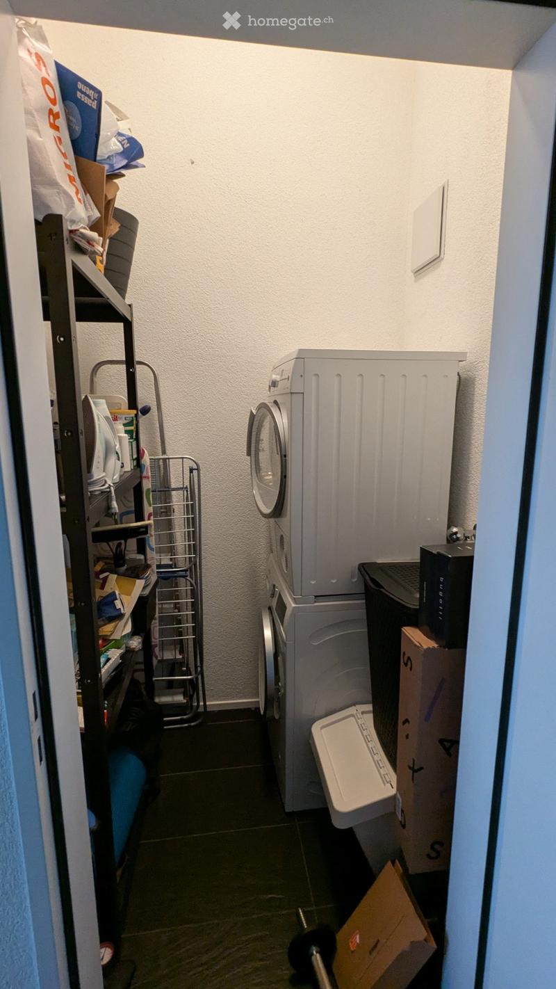 A small utility room with a washing machine, dryer, shelf with cleaning supplies, ironing board, and a cart.