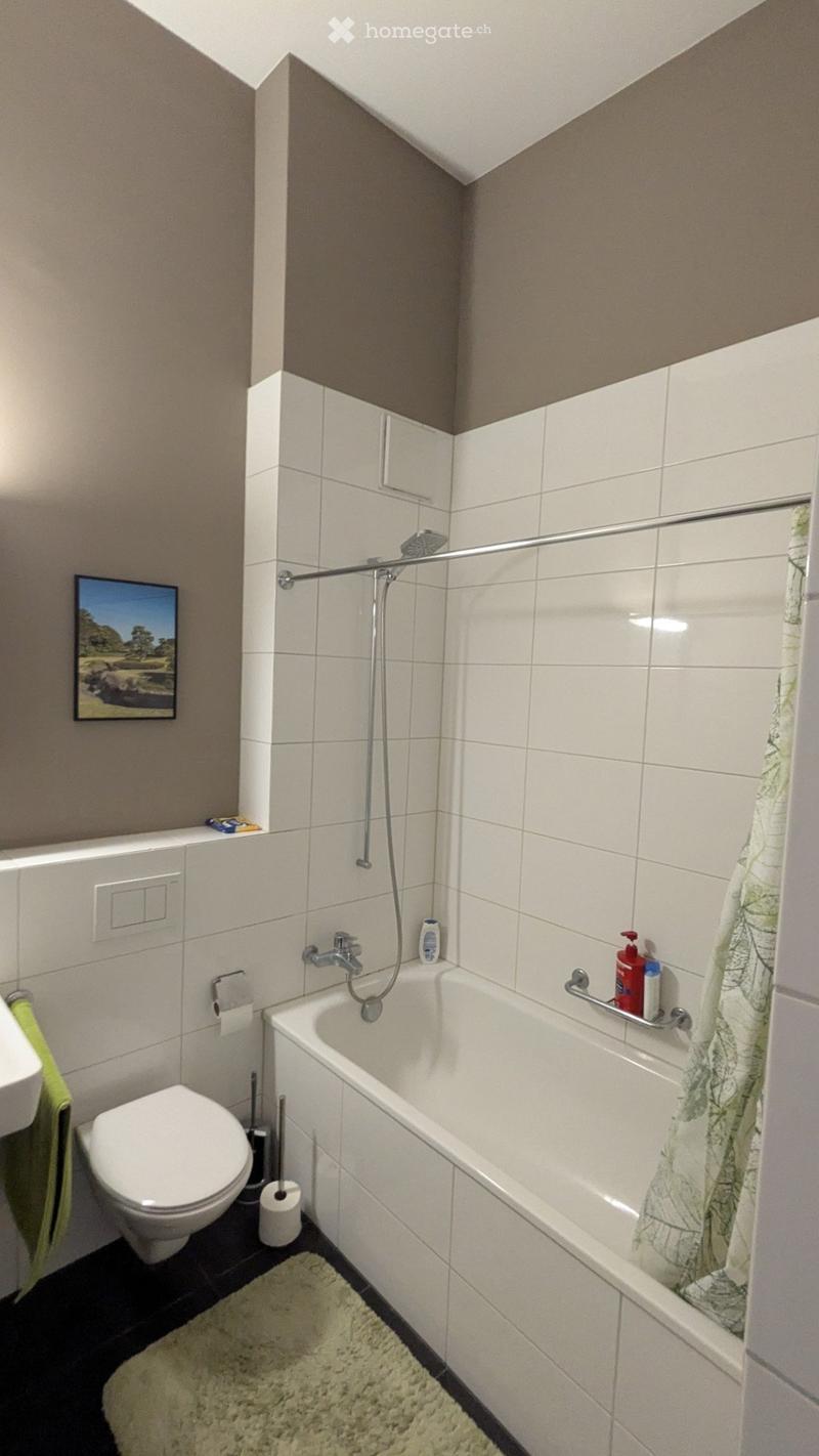Modern bathroom with white tiles, bathtub, toilet, showerhead, towel, and a picture frame on the wall.